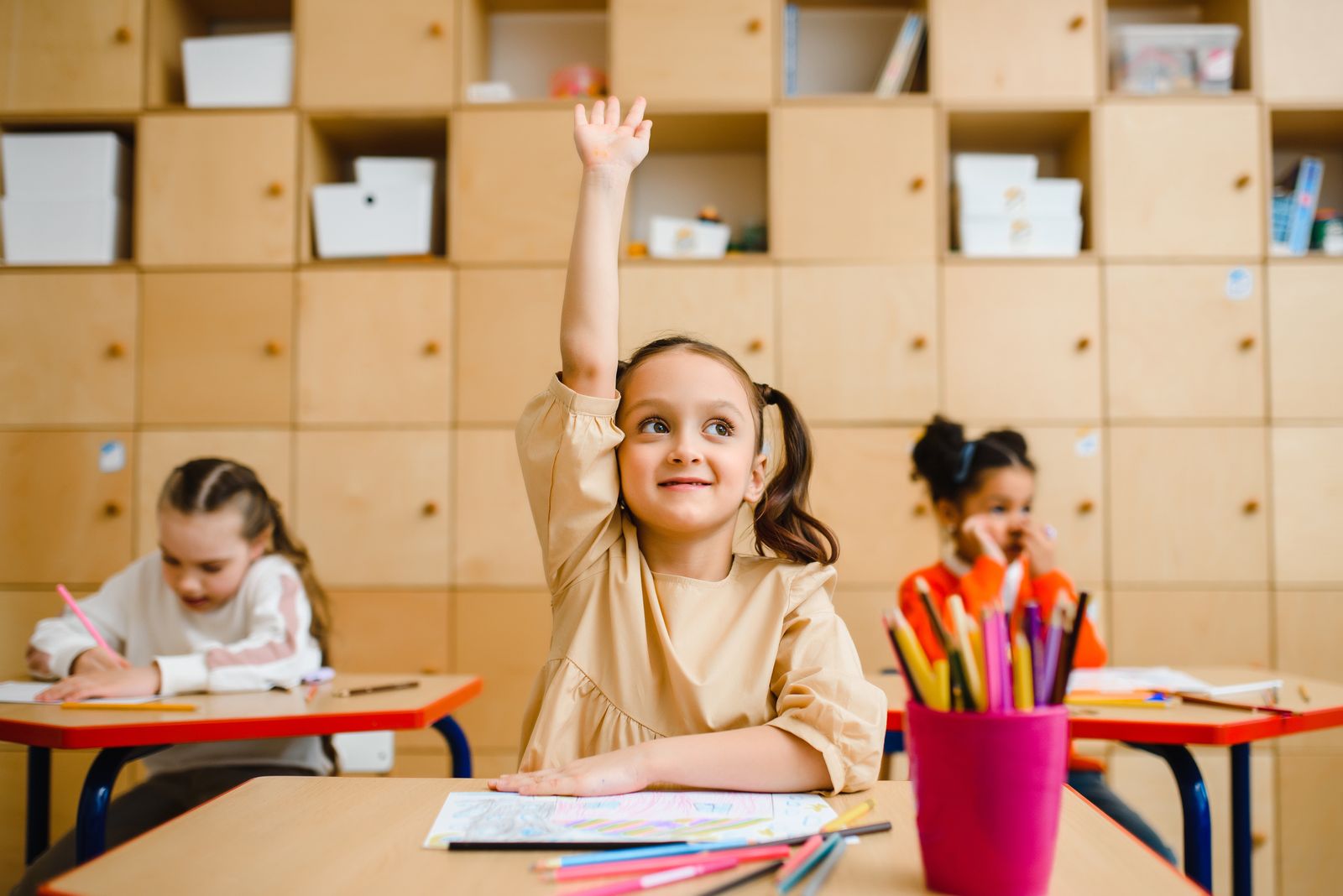 Smiling girl with pigtails raising her hand at her desk in a bright classroom with classmates nearby