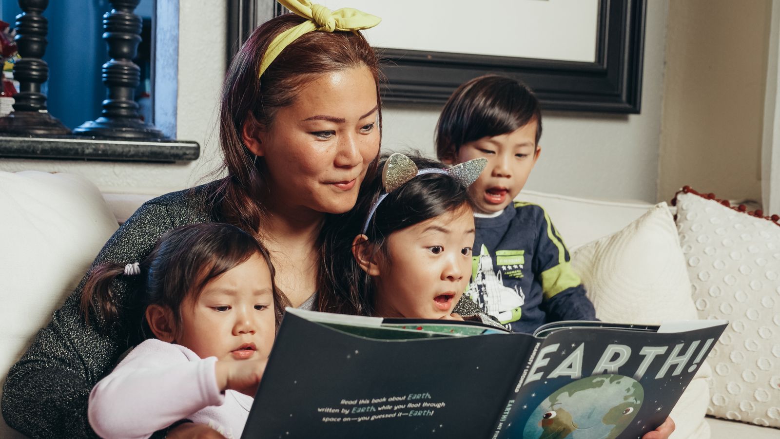Mother sitting on a couch reading a picture book aloud to three young children gathered around her