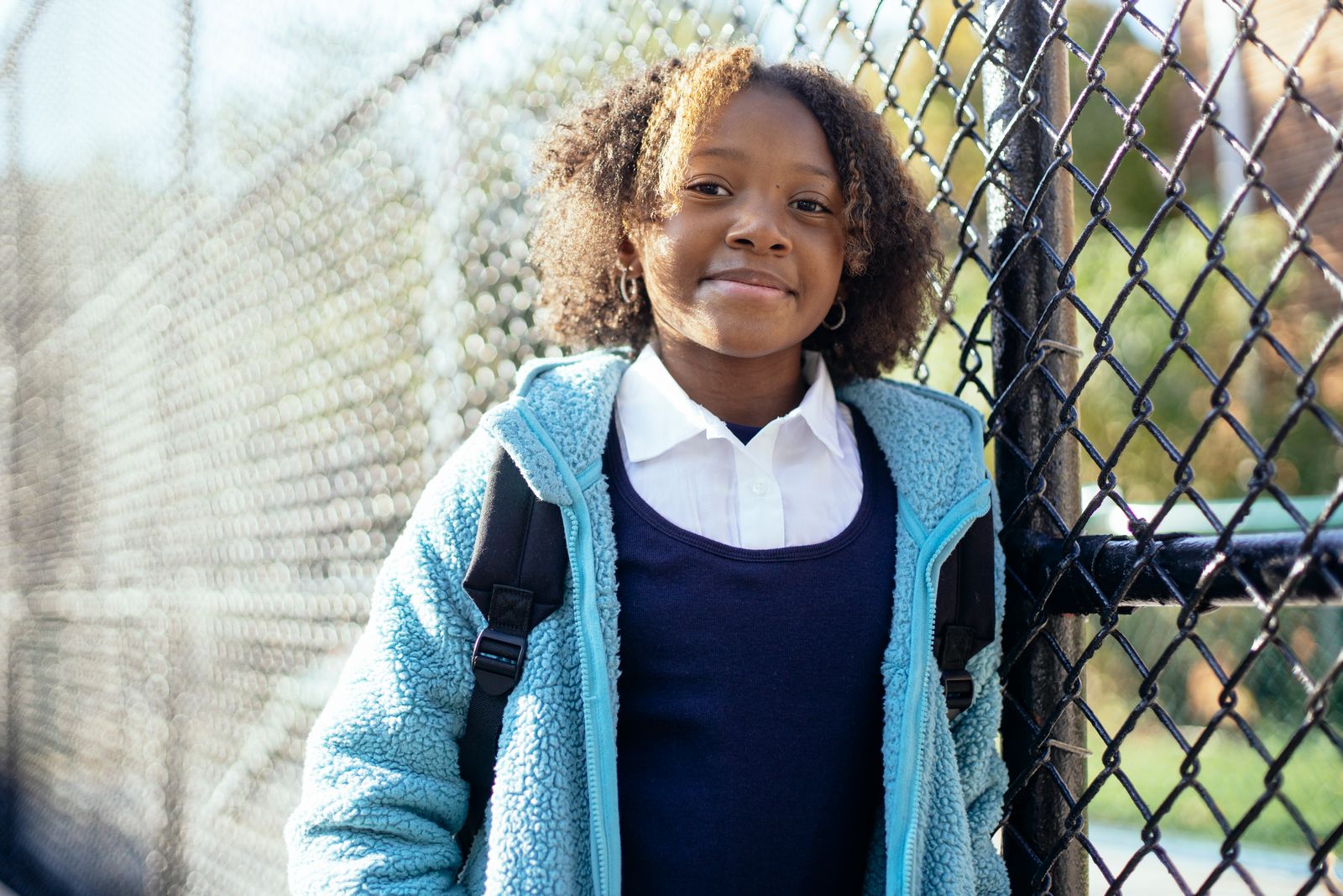 Smiling girl in a school uniform and blue jacket standing by a chain-link fence in her neighborhood