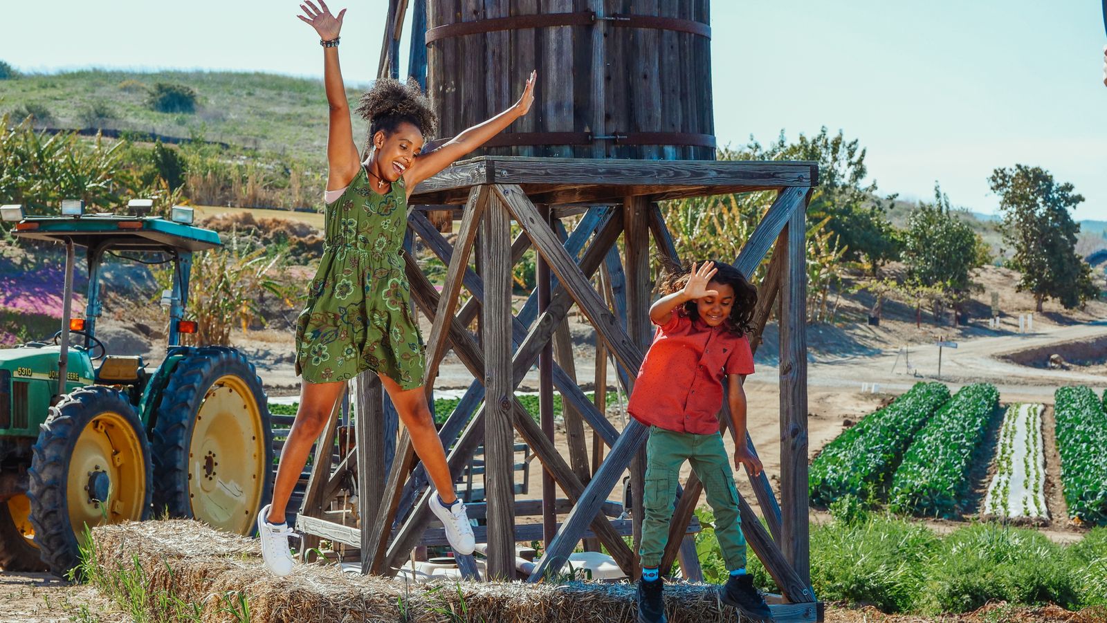 Two children playing on a wooden farm platform beside a green tractor and rows of crops
