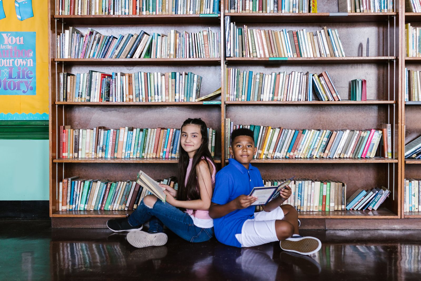 Two students sitting on the floor in front of tall bookshelves reading books