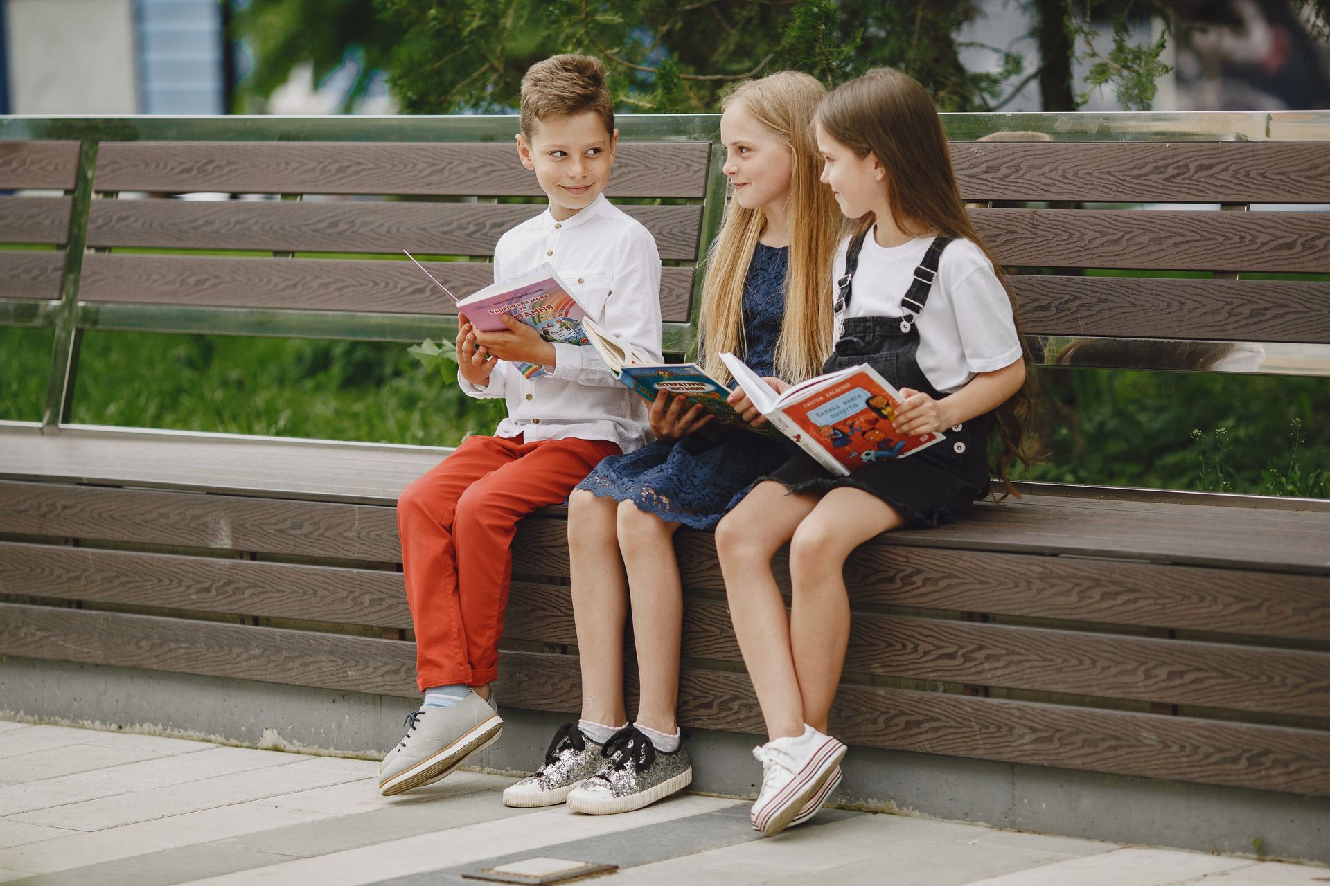 Children sitting on a wooden bench outdoors, each reading a book