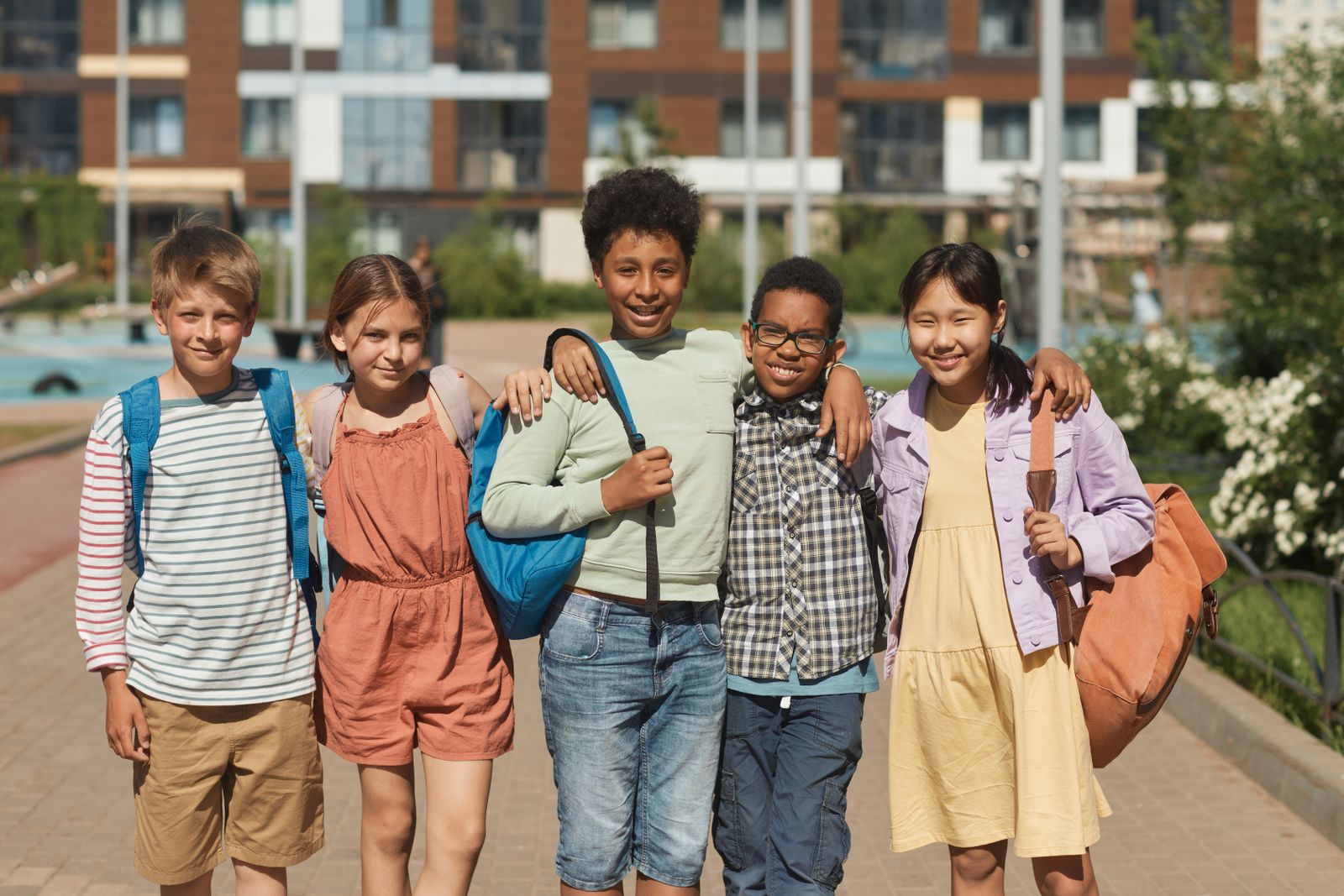 Group of smiling elementary students with backpacks standing together on a city sidewalk in front of apartment buildings